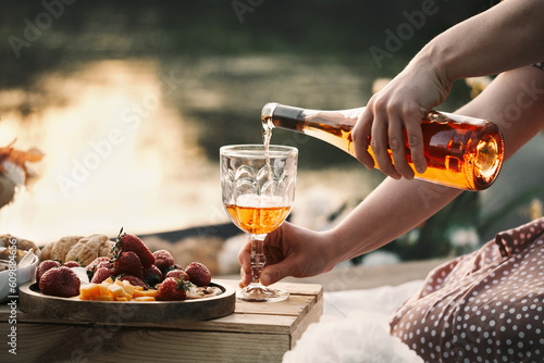 Rose wine on sunset picnic outdoors in summer. Glass of drink, fresh strawberries, cheese and pie for romantic eco style dinner. Woman pouring drink from the bottle 