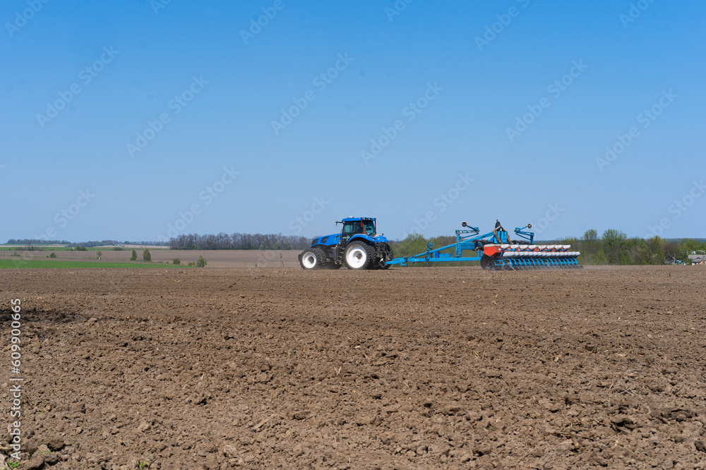 Obraz premium The tractor drives across the field and cultivates the land. Agricultural vehicle works in countryside. Sowing is the process of planting seeds in the ground as part of the early spring time
