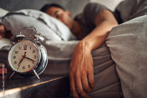 Alarm clock, relax and man sleeping in the bed of his modern apartment in the morning. Lazy, resting and closeup of a timer bell with a male person taking a nap and dreaming in bedroom at his home. photo