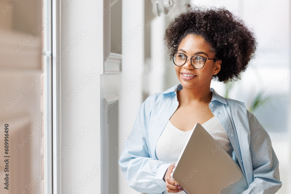 © JenkoAtaman - African american business woman standing with laptop near window in modern office