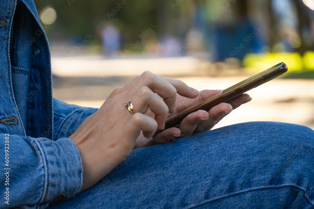 Close-up of a woman's hands holding a smartphone. Young woman in jeans and a denim jacket sitting on a park bench reads a text and watches a video on her mobile phone.