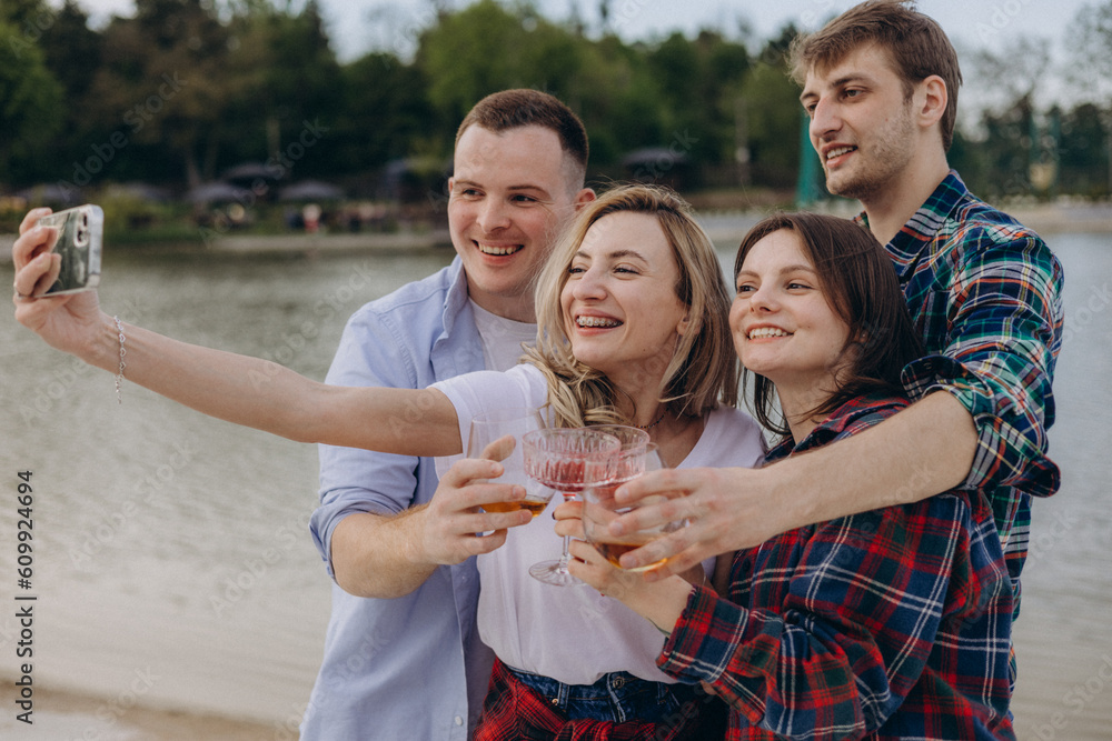 Obraz premium Stylish young people in casual clothes waving hand, posing for smartphone camera when taking selfie or shooting video for social media using mobile phone by lake. Friends having rest and fun.