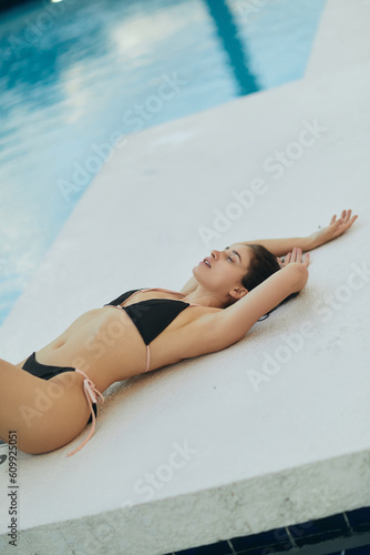 stunning figure, sun-kissed woman in black bikini, sexy model with wet hair laying down while posing next to swimming pool in luxury resort, Miami, Florida, USA, blurred background