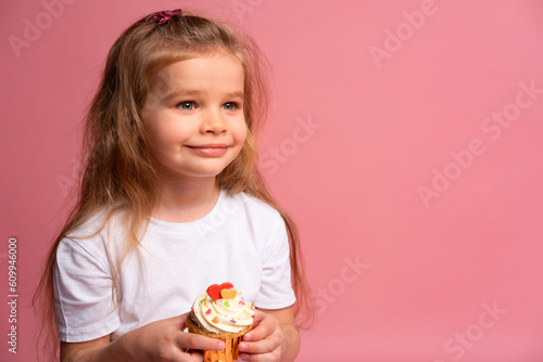 Happy little girl holds birthday cupcake makes wish on pink background
