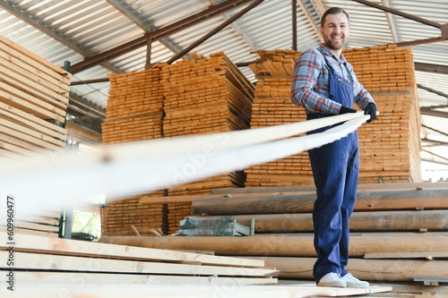 Portrait of a handsome worker choosing the best wooden boards. Carpenter standing next to a big stack of wood bars in a warehouse.