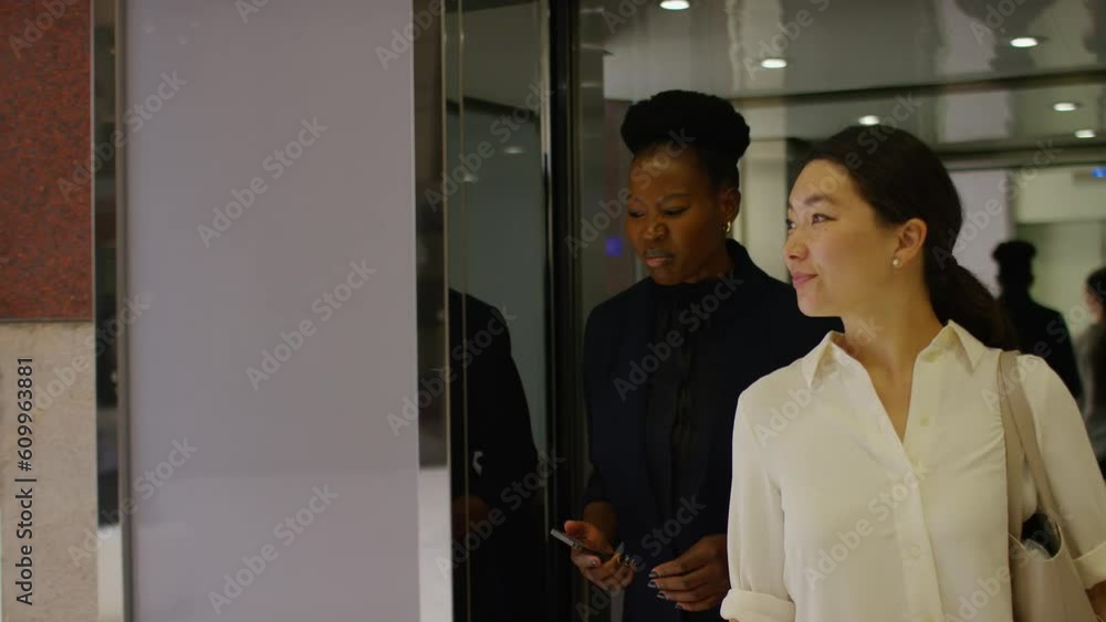 Two female colleagues exit an elevator and talk as they walk through a building lobby, in slow motion Two female colleagues exit an elevator and talk as they walk through a building lobby, in slow motion