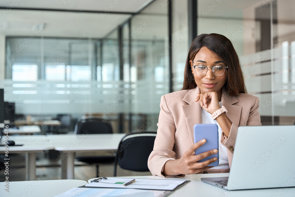 Young busy professional African American business woman executive manager holding cellphone using mobile looking at cell phone tech sitting at desk in office working on laptop, copy space. Young busy professional African American business woman executive manager holding cellphone using mobile looking at cell phone tech sitting at desk in office working on laptop, copy space.