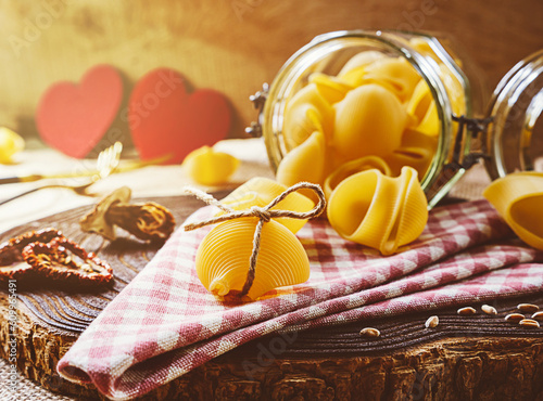 Lumaconi pasta in a glass jar on a wooden background in the morning sun. White mushrooms, tomatoes, olive oil, wheat and two red hearts. Valentine's Day. photo