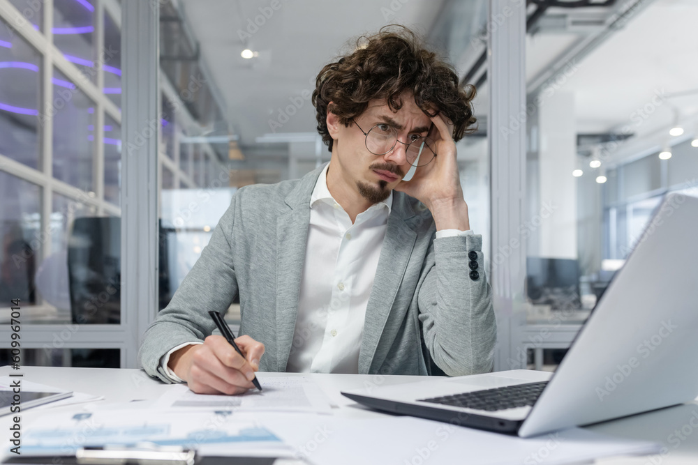 Upset businessman behind paper work inside modern office, mature man with beard reading financial reports and account documents unhappy with results and disappointed with achievements
