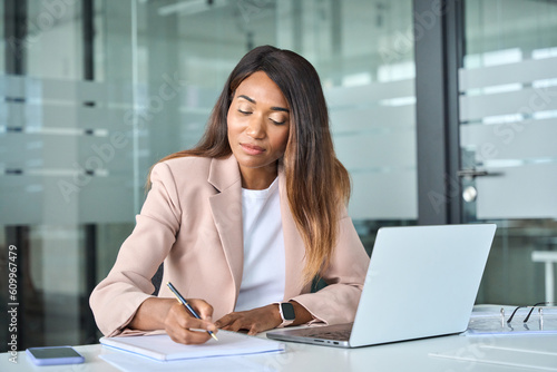 Busy professional young African American business woman company manager, female executive or secretary wearing suit working in office with laptop computer writing notes sitting at desk.