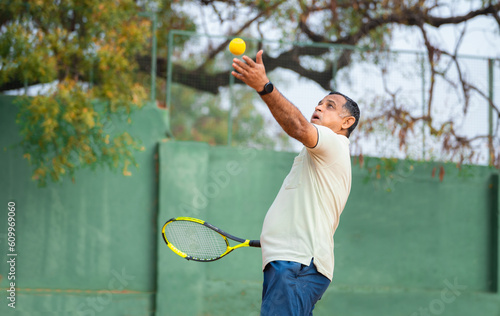 Indian senior man servicing by throwing ball at tennis court while playing game - concept of tournament, competition and fitness. © WESTOCK