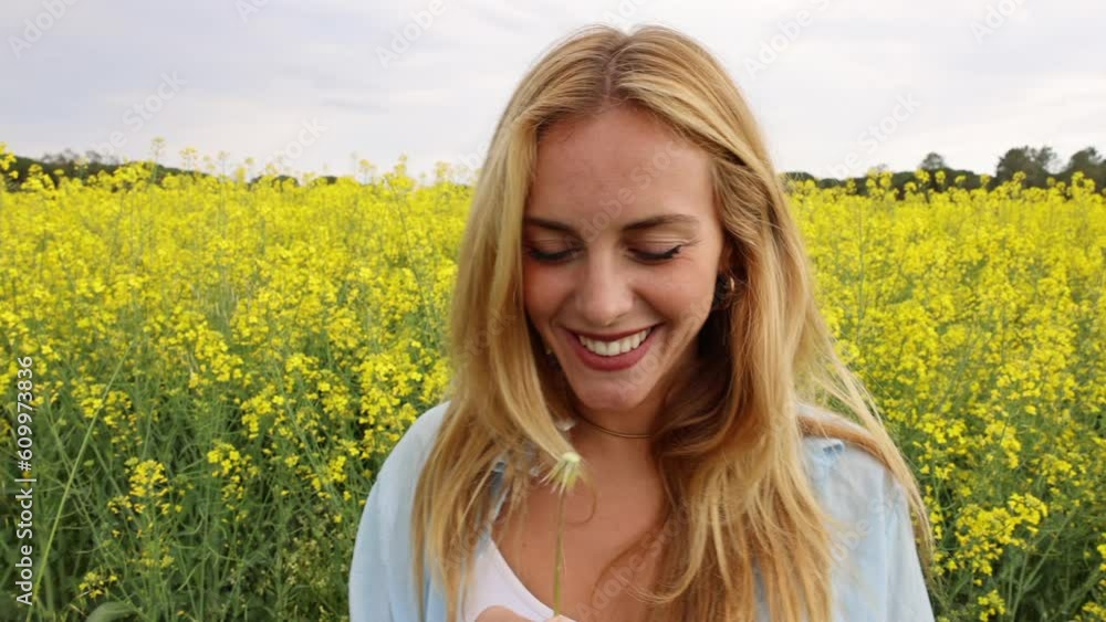 Beautiful young blonde cheerful woman, blowing dandelion seeds amidst a field of blooming yellow rapeseed flowers Beautiful young blonde cheerful woman, blowing dandelion seeds amidst a field of blooming yellow rapeseed flowers