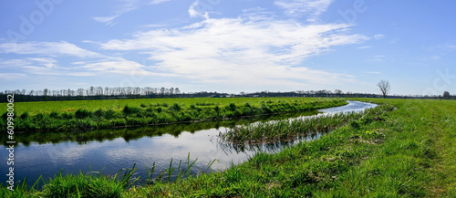 Panorama photo with bright colors and lots of sunlight, of a green grass landscape with a river with reflection. In a blue sky with white clouds.