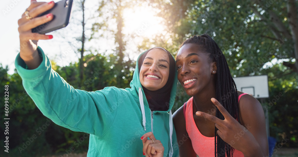 Fototapeta premium Portrait of Two Multiethnic Female Friends in Sports Clothes Posing for a Selfie Using a Smartphone. Young Women Celebrating a Win on an Outdoor Basketball Court by Taking a Photo for Social Media