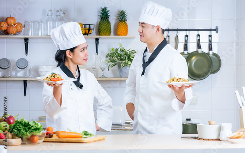 Two Asian male and female professional chef wearing uniform, holding plate, presenting of cooking competition, looking each other, standing in kitchen.