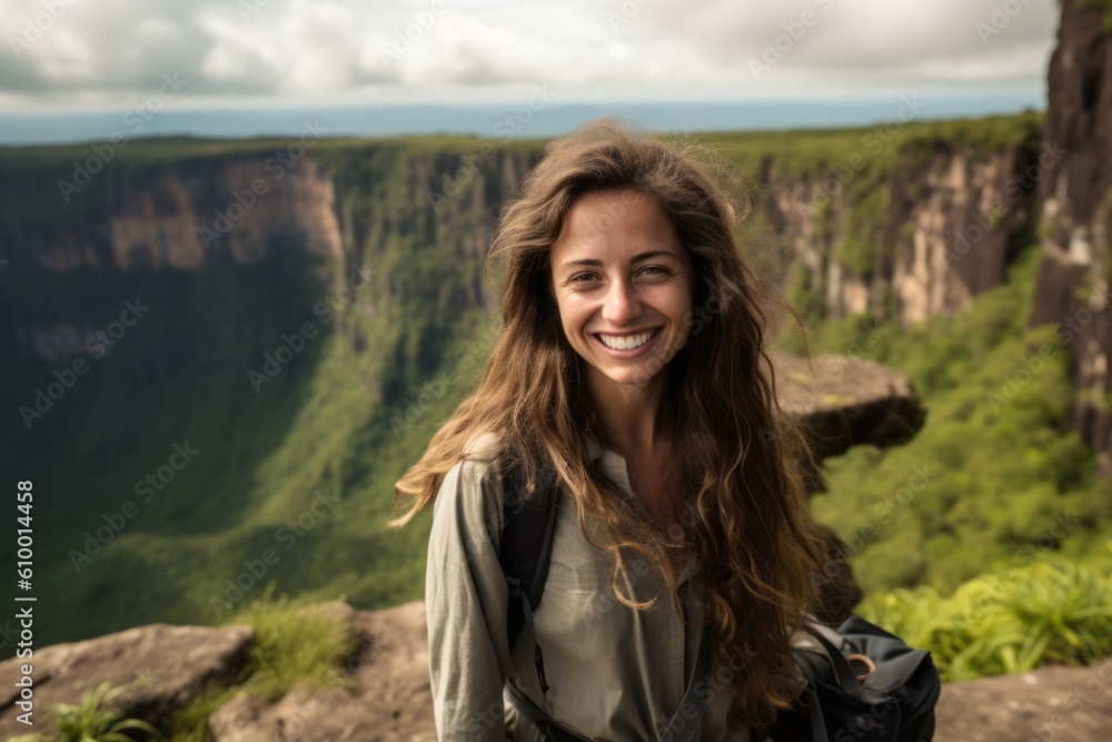 Beautiful young woman hiker with backpack standing on top of a cliff and looking at camera.