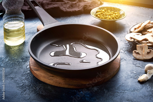 Dark frying pan with poured oil on a dark blue abstract background. Nearby is a plate of champignons, green peas, and a bottle of oil.