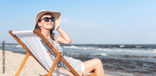 Happy brunette woman wearing sunglasses and hat relaxing on a wooden deck chair at the ocean beach.
