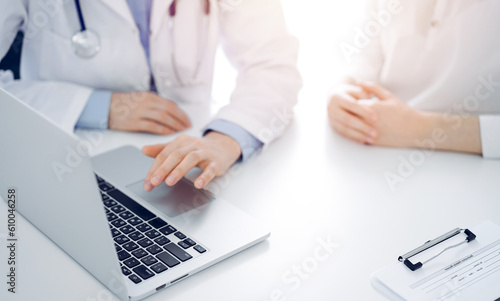 Doctor and patient sitting near each other at the desk in clinic. The focus is on female physician s hands using laptop computer  close up. Medicine concept.