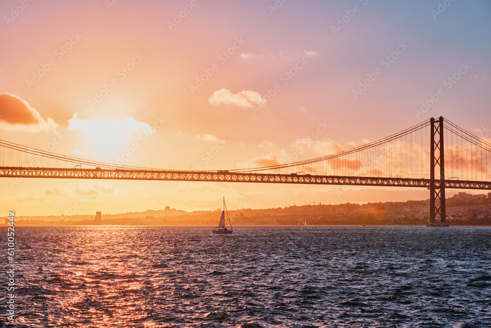 Obraz premium View of 25 de Abril Bridge famous tourist landmark of Lisbon connecting Lisboa and Almada on Setubal Peninsula over Tagus river with tourist yacht silhouette at sunset. Lisbon, Portugal