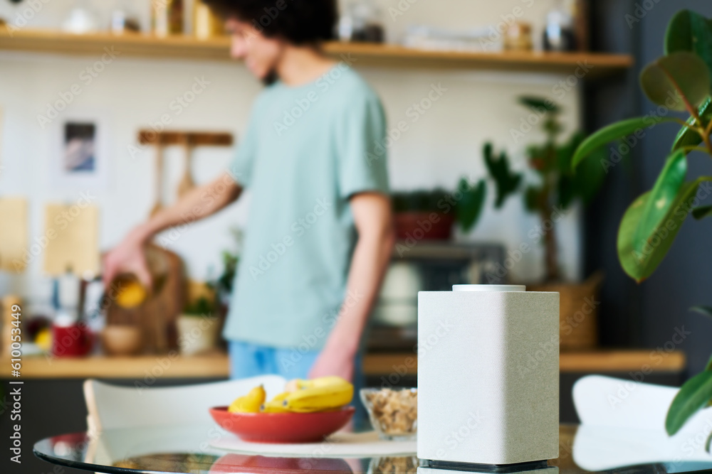 Close-up of smart speaker on kitchen table to listen to music or news with man preparing breakfast in background Close-up of smart speaker on kitchen table to listen to music or news with man preparing breakfast in background