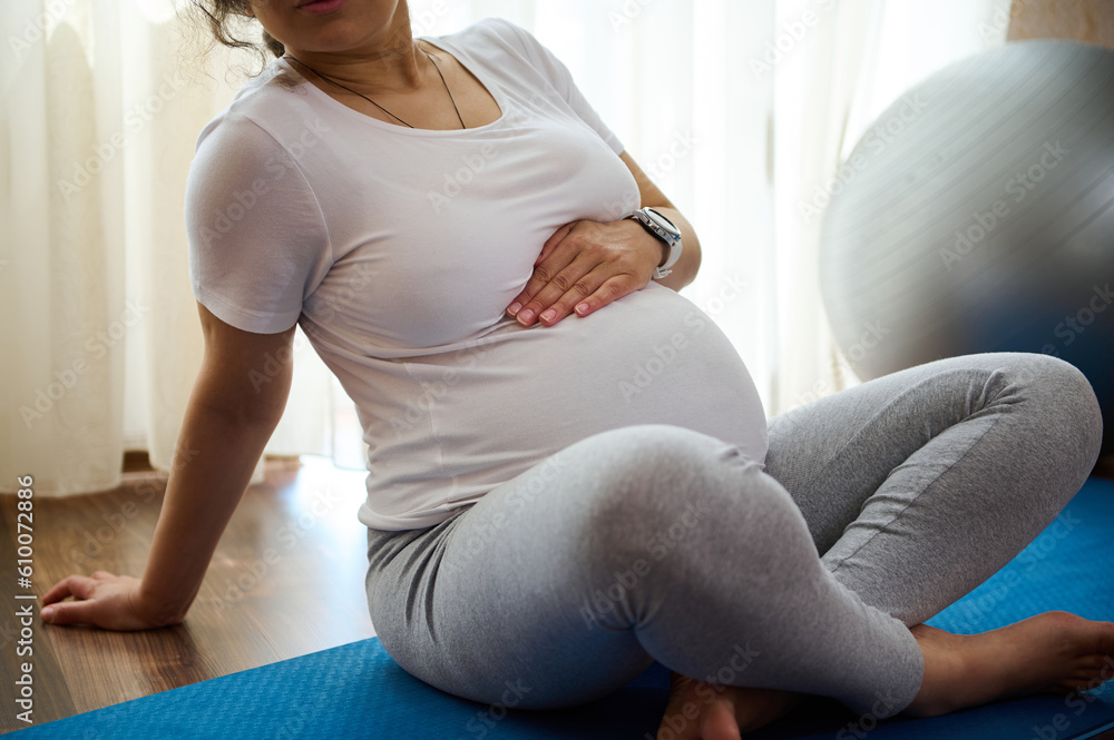 Close-up pregnant millennial woman sitting on exercise mat, touches her belly after performing prenatal and meditation exercises at cozy tranquil homely atmosphere. Pregnancy and maternity lifestyle Close-up pregnant millennial woman sitting on exercise mat, touches her belly after performing prenatal and meditation exercises at cozy tranquil homely atmosphere. Pregnancy and maternity lifestyle