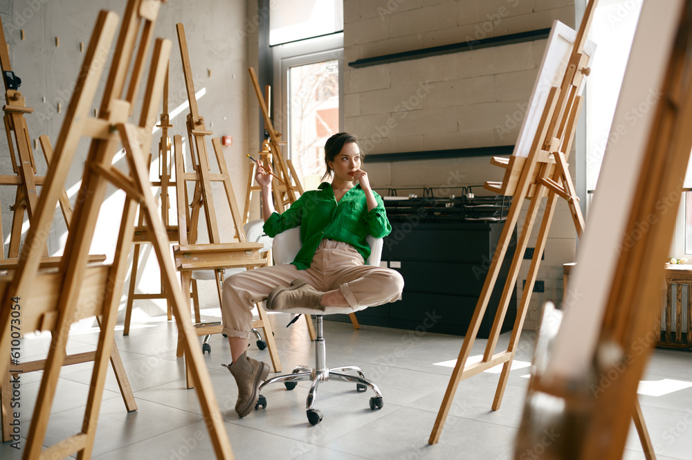 Obraz premium Female artist with paintbrush in hand looking seriously on her own picture