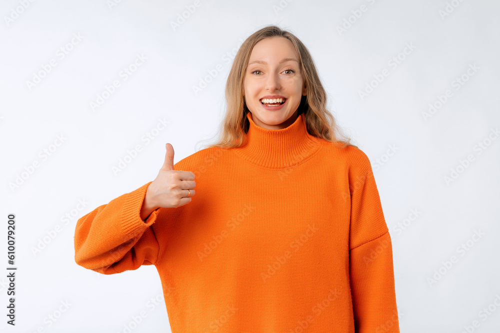 Lovely happy smiling young woman shows thumb up, recommend and approve something good, wears soft orange sweater, standing over neutral studio background Lovely happy smiling young woman shows thumb up, recommend and approve something good, wears soft orange sweater, standing over neutral studio background