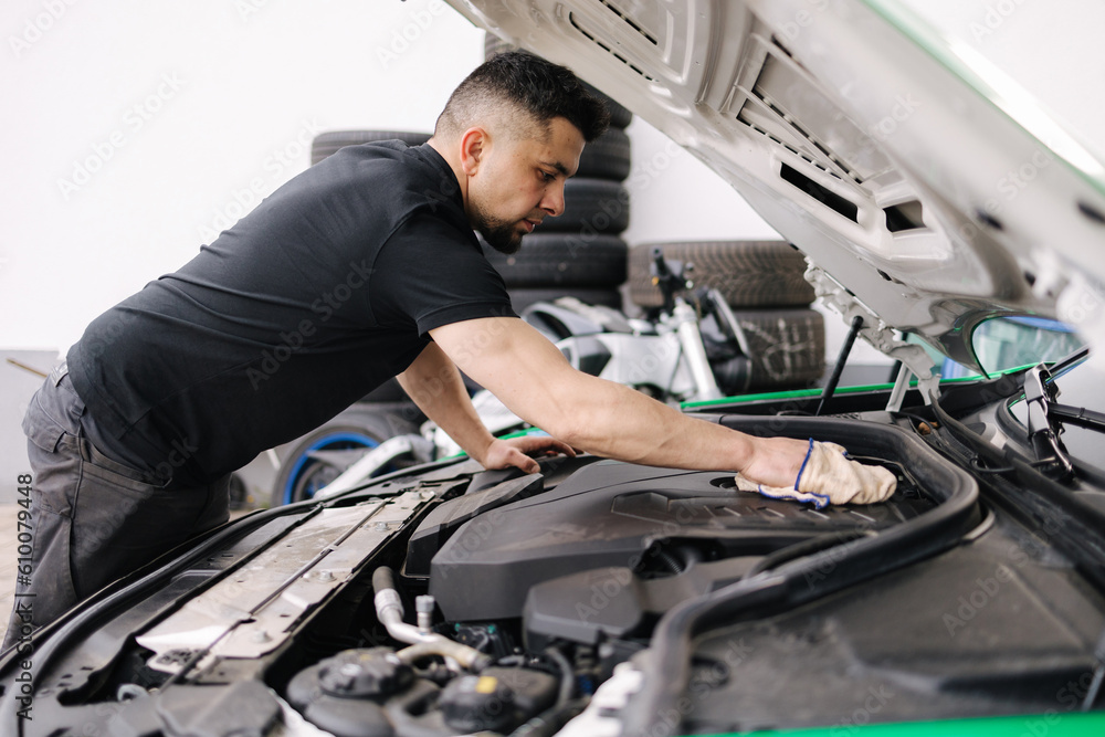 Side view of a male mechanic working in his garage. Young bearded man checks the engine in the car, the hood is open Side view of a male mechanic working in his garage. Young bearded man checks the engine in the car, the hood is open