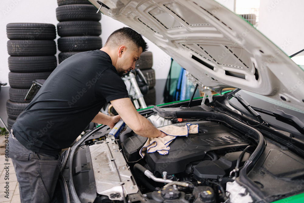 Side view of a male mechanic working in his garage. Young bearded man checks the engine in the car, the hood is open Side view of a male mechanic working in his garage. Young bearded man checks the engine in the car, the hood is open