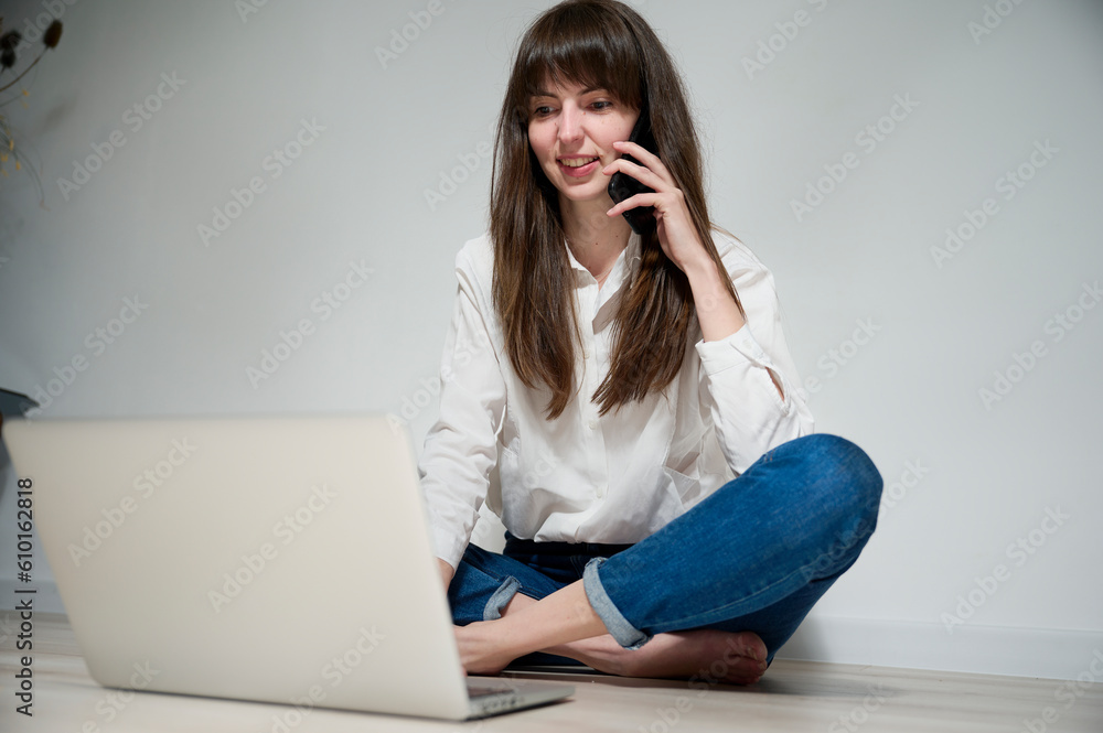 An attractive girl sitting on the floor works remotely on her laptop, responding to various messages and browsing the internet. Freelance work, remote learning, project work.