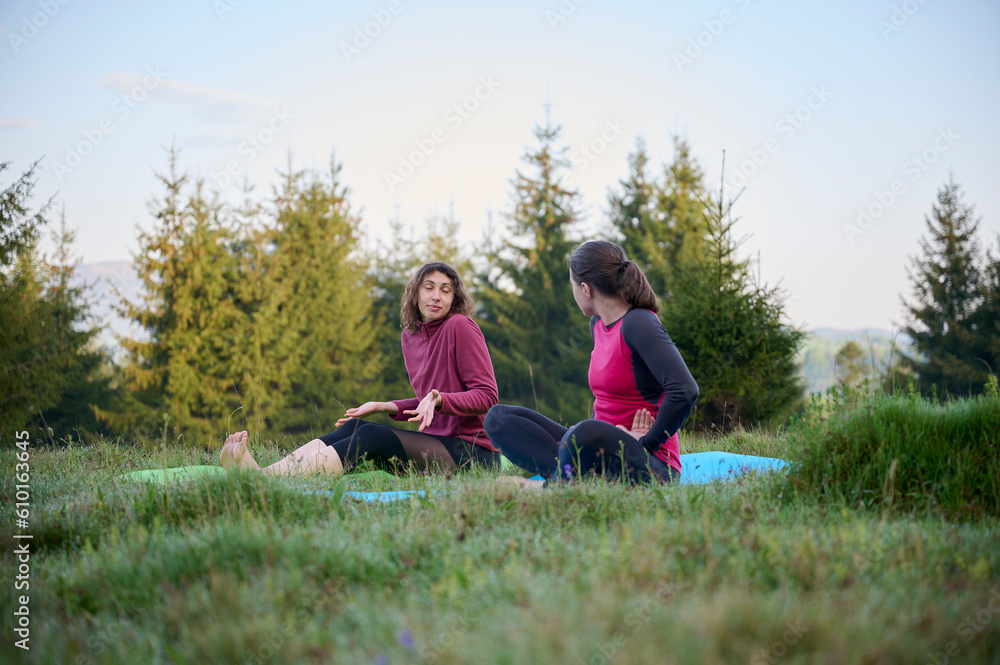 Fototapeta premium Two athletic young women dressed in sporty attire engage in a range of yoga poses amidst the scenic beauty of mountainous surroundings. The gentle rays of the morning sun cast on their slender bodies