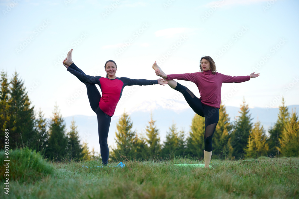 Two athletic young women dressed in sporty attire engage in a range of yoga poses amidst the scenic beauty of mountainous surroundings. The gentle rays of the morning sun cast on their slender bodies