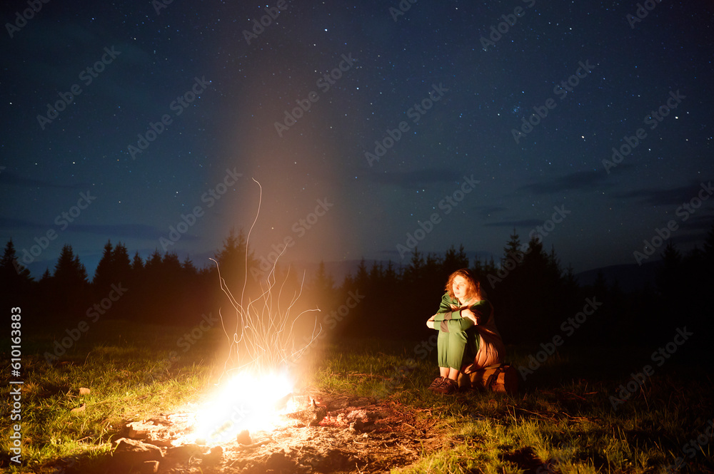 A young female traveler sits by the campfire near her tent, enjoying a moment of relaxation beneath the beautifully starry sky. As the crackling flames provide warmth and comfort A young female traveler sits by the campfire near her tent, enjoying a moment of relaxation beneath the beautifully starry sky. As the crackling flames provide warmth and comfort