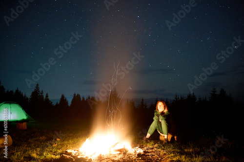 A young female traveler sits by the campfire near her tent, enjoying a moment of relaxation beneath the beautifully starry sky. As the crackling flames provide warmth and comfort © Василь Івасюк