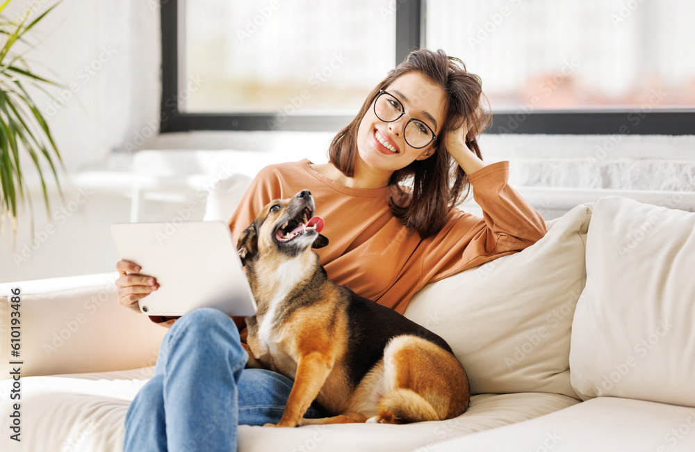 Happy young woman spending time with her pet dog at home sitting on couch and browsing in tablet Happy young woman spending time with her pet dog at home sitting on couch and browsing in tablet