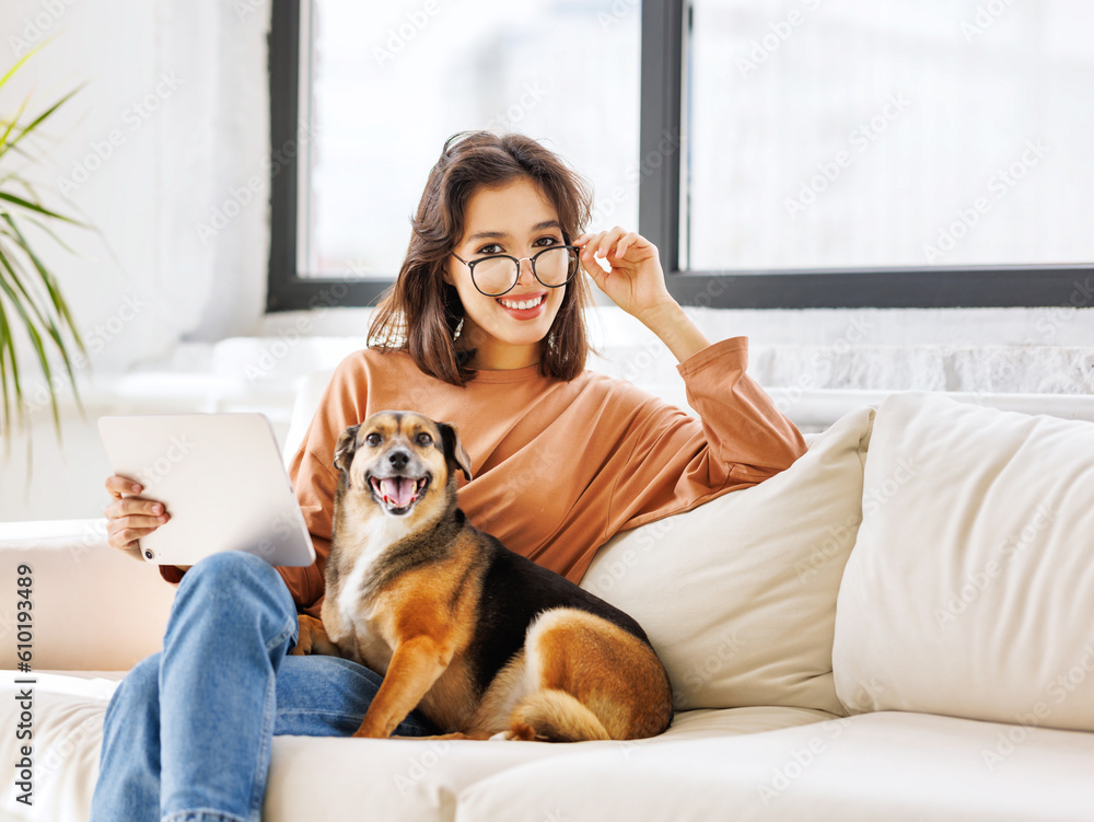 Happy young woman spending time with her pet dog at home sitting on couch and browsing in tablet Happy young woman spending time with her pet dog at home sitting on couch and browsing in tablet
