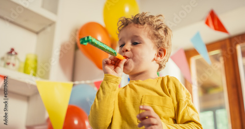 Low Angle Portrait of a Male Kid Using a Party Blower in a Living Room Decorated by Balloons. Little Cute Boy Blowing a Party Horn and Laughing on his Birthday. Happy Toddler Celebrating and Smiling  © Kitreel