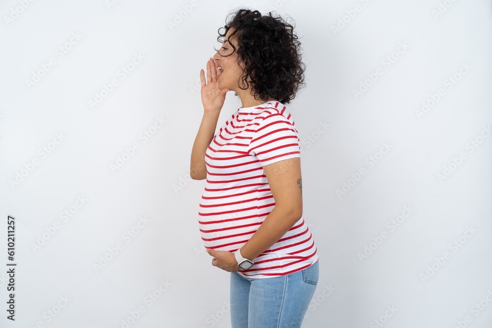Half-faced close up portrait of astonished amazed young pregnant woman wearing striped t-shirt over white background holding hand near mouth Half-faced close up portrait of astonished amazed young pregnant woman wearing striped t-shirt over white background holding hand near mouth