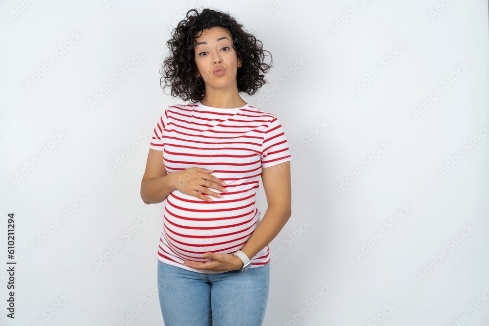 Shot of pleasant looking young pregnant woman wearing striped t-shirt over white background , pouts lips, looks at camera, Human facial expressions Shot of pleasant looking young pregnant woman wearing striped t-shirt over white background , pouts lips, looks at camera, Human facial expressions
