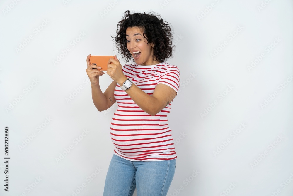 Portrait of an excited young pregnant woman wearing striped t-shirt over white background playing games on mobile phone. Portrait of an excited young pregnant woman wearing striped t-shirt over white background playing games on mobile phone.