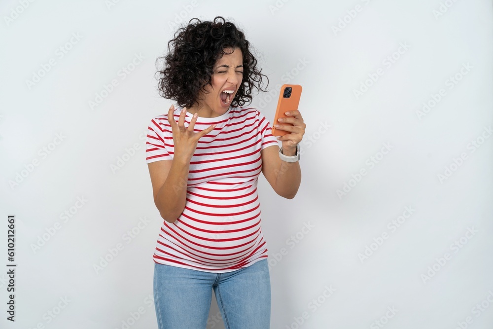 Angry young pregnant woman wearing striped t-shirt over white background screaming on the phone, having an argument with an employee. Troubles at work. Angry young pregnant woman wearing striped t-shirt over white background screaming on the phone, having an argument with an employee. Troubles at work.