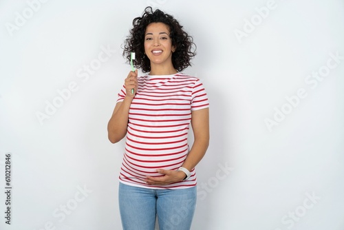 young pregnant woman wearing striped t-shirt over white background holding a toothbrush and smiling. Dental healthcare concept.
