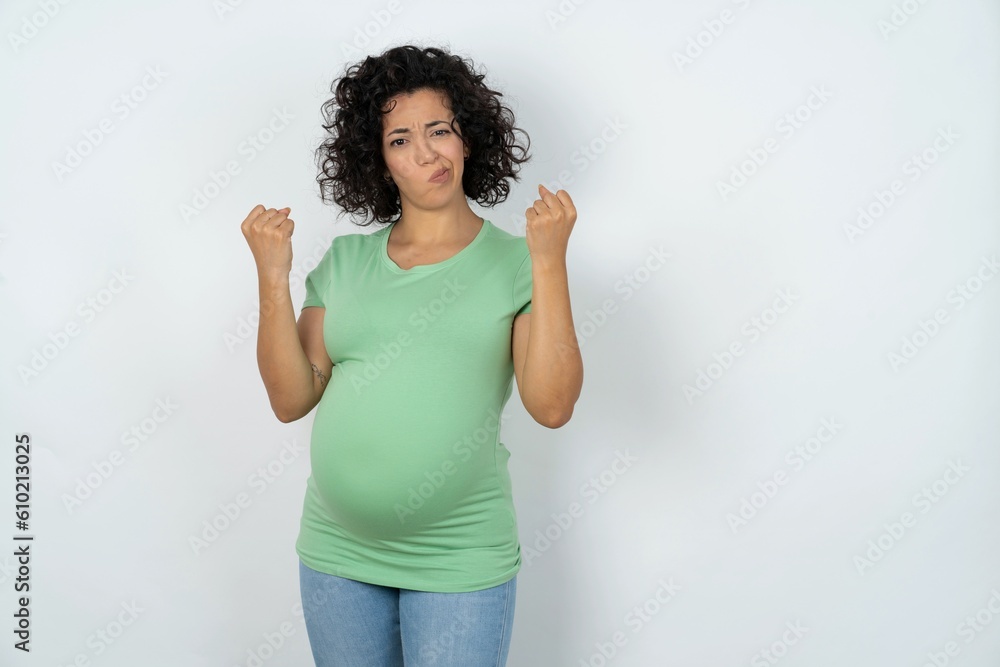 Irritated young pregnant woman wearing green t-shirt over white background blows cheeks with anger and raises clenched fists expresses rage and aggressive emotions. Furious model Irritated young pregnant woman wearing green t-shirt over white background blows cheeks with anger and raises clenched fists expresses rage and aggressive emotions. Furious model