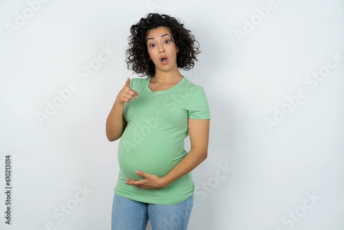 Shocked young pregnant woman wearing green t-shirt over white background points at you with stunned expression