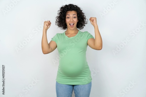 Shocked ecstatic young pregnant woman wearing green t-shirt over white background win luck lottery raise hands up shout yea