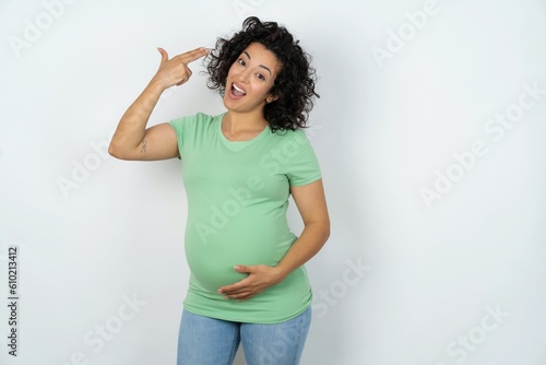 Quadro su tela young pregnant woman wearing green t-shirt over white background foolishness around shoots in temple with fingers makes suicide gesture