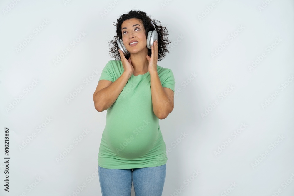 Joyful young pregnant woman wearing green t-shirt over white background sings song keeps hand near mouth as if microphone listens favorite playlist via headphones Joyful young pregnant woman wearing green t-shirt over white background sings song keeps hand near mouth as if microphone listens favorite playlist via headphones