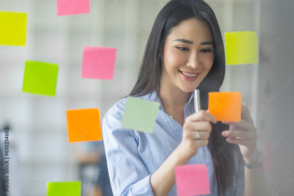 Smiling business Asian people thinking and use sticky notes on glass wall in office with her colleague in background.
Smiling business Asian people thinking and use sticky notes on glass wall in office with her colleague in background.