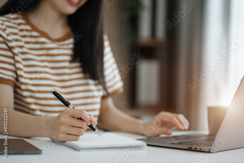 woman sitting at a table at home working on a laptop and writing down ideas in a notebook.